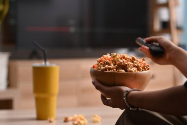 Evde televizyon izlerken elinde patlamış mısır ve uzaktan kumanda tutan genç bir kadın..