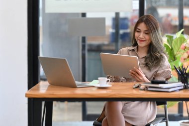 Modern işyerinde otururken dijital tablet kullanan çekici bir iş kadını..