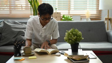 Genç adam oturma odasında gözlük takıp kitap okuyor..