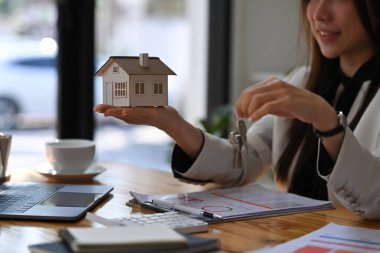 Evin modelini ve anahtarlarını elinde tutan gülümseyen kadın. Gayrimenkul yatırımı ve sigorta kavramı.