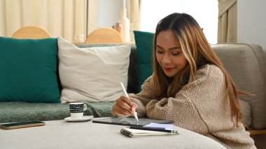 Güzel Asyalı kadın oturma odasında oturuyor ve dijital tabletle internette çalışıyor..