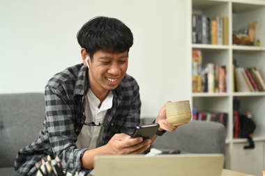 Genç adam kahve içiyor ve evde akıllı telefon kullanıyor..