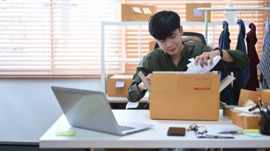 Genç adam çevrimiçi satıcı dizüstü bilgisayardan online siparişleri kontrol ediyor ve müşterilere teslimat için paketler hazırlıyor.