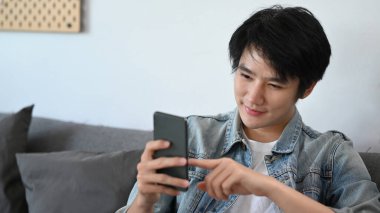 Mutlu Asyalı adam kanepede oturup mesaj okuyor ya da akıllı telefondan sosyal medyayı kontrol ediyor..