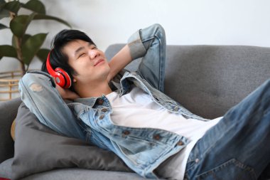 Mutlu Asyalı adam kablosuz kulaklıkla müzik dinliyor ve evdeki kanepede uzanıyor..