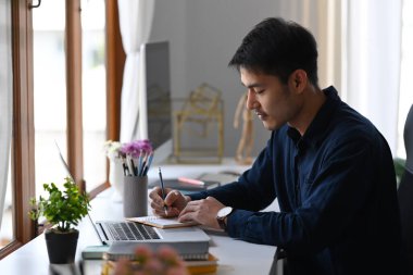 Dizüstü bilgisayarla çalışan ve not defterine not alan konsantre Asyalı adam..