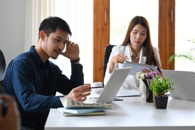 Ciddi Asyalı ofis çalışanı ofiste meslektaşıyla otururken dizüstü bilgisayara odaklanmış görünüyor..