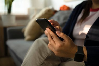 Evdeki kanepede otururken akıllı telefondan kısa mesaj okuyan kısa boylu bir kadın..