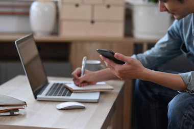 Akıllı telefonu tutan ve bir deftere fikirler yazan genç bir adam..