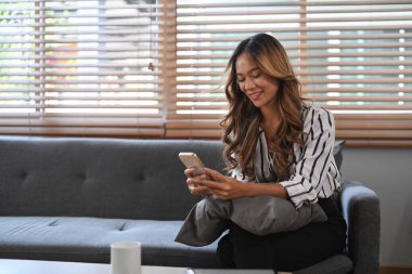 Evdeki koltukta oturan ve cep telefonu kullanan gülümseyen genç kadın..