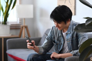 Rahat Asyalı bir adam, gündüz vakti evdeki koltukta otururken cep telefonu kullanıyor..