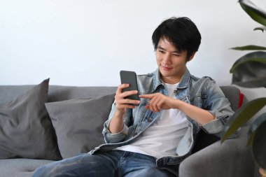 Sıradan giyinen mutlu adam akıllı telefon kullanıyor, sosyal medyayı kontrol ediyor, koltukta dinlenirken sipariş veriyor..