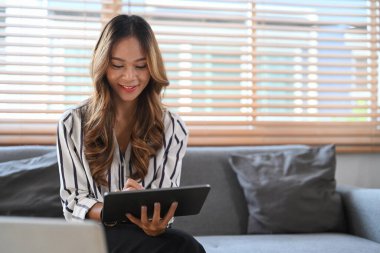 Mutlu genç bir kadın evdeki kanepede dinleniyor ve dijital tabletteki sosyal medya paylaşımlarını kontrol ediyor..