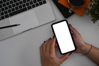 Yukarıda, ofis masasında boş ekranlı cep telefonu tutan genç bir kadın var..