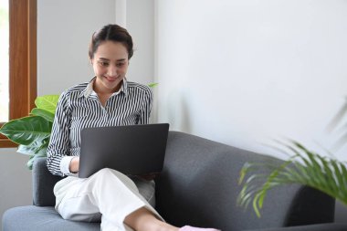 Gülümseyen genç kadın kanepede oturuyor ve evinde dizüstü bilgisayarla serbest proje yapıyor..