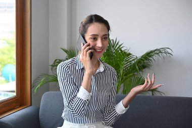 Memnun genç iş kadını koltukta oturuyor ve cep telefonuyla konuşuyor..