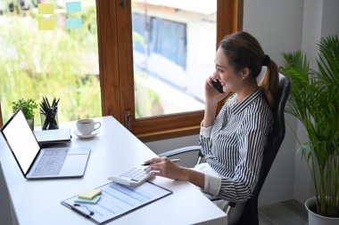 Genç çekici bayan yönetici modern ofiste oturuyor ve cep telefonuyla konuşuyor..