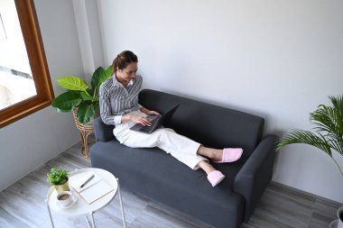 Parlak oturma odasındaki kanepede dinlenen ve dizüstü bilgisayar kullanan genç bir kadın manzarası..