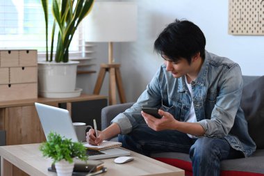 Bilgisayarla çalışan ve oturma odasında cep telefonu kullanan memnun bir adam..