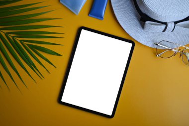 Dijital tablet, şapka, güneş kremi şişeleri ve sarı arka planda yeşil yaprak palmiyesi. Seyahat, yaz ve tatil konsepti.
