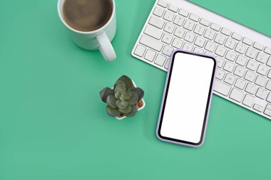 Boş ekranlı akıllı telefon, kahve fincanı ve yeşil arka planda kablosuz klavye. Üst görünüm.