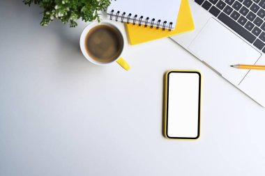 Üzerinde cep telefonu, laptop, kahve fincanı, kırtasiye ve saksı bitkisi olan beyaz ofis masası..