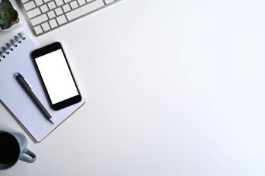 Üst görünüm cep telefonu, defter, kahve fincanı ve beyaz ofis masasındaki kablosuz klavye..