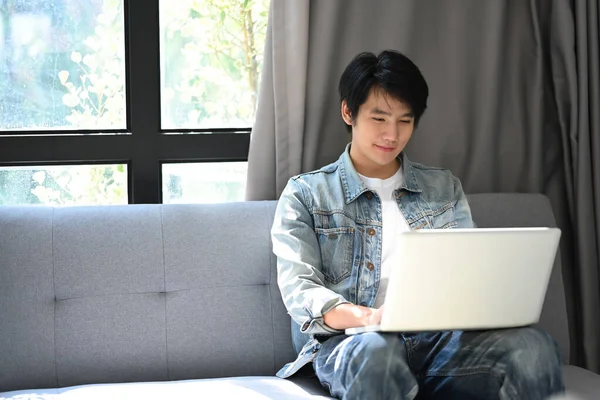 カジュアルなアジアの男は自宅でソファに休んで コンピュータのラップトップでインターネットを閲覧 — ストック写真