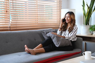 Güzel Asyalı kadın kanepede oturuyor ve cep telefonuyla konuşuyor, evde vakit geçiriyor..