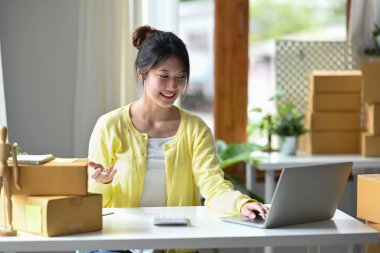 Smiling young female entrepreneur checking online order on digital tablet and preparing parcel boxes for delivery to customers