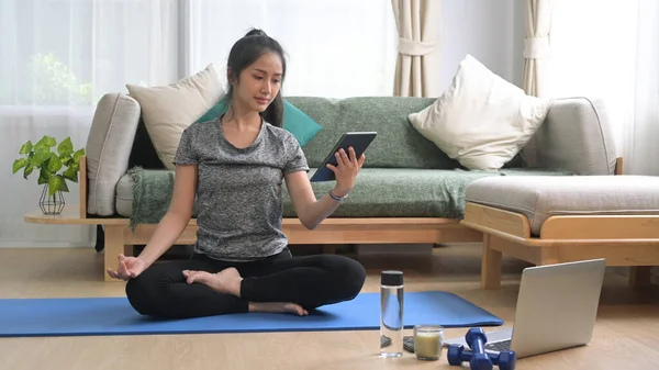 Hermosa joven sentada en una esterilla de yoga y viendo clases de fitness en línea en una tableta digital — Foto de Stock