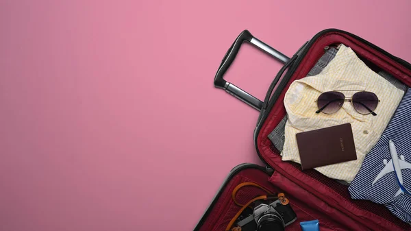 Otwarta walizka z okularami przeciwsłonecznymi, kamerą, paszportem i ubraniami na różowym tle. Przygotowanie do wakacji letnich — Zdjęcie stockowe