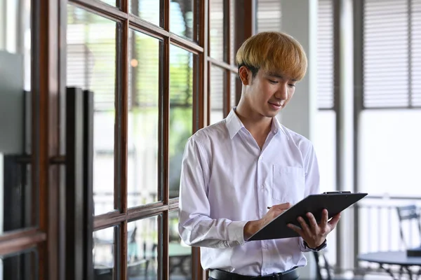 Asian male office worker reading information on clipboard while standing at modern office — Foto de Stock