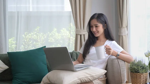 Casual young asian woman resting on couch and using laptop, surfing internet, checking email — Zdjęcie stockowe