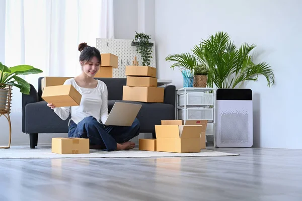 Happy female online seller checking online order on laptop and preparing packages for delivery to customers