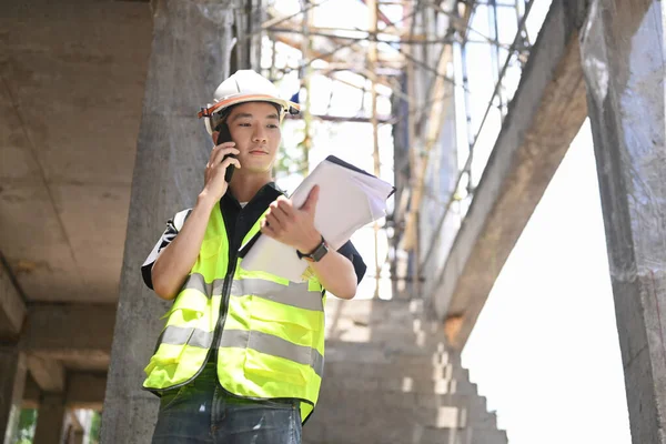 Young engineer man in helmet talking on mobile phone under new building — Stock Photo, Image