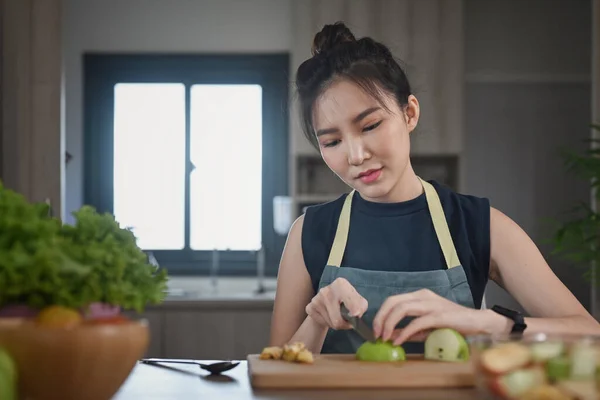 Mulher asiática em avental sentado na mesa da cozinha e maçã de corte na placa de corte de madeira durante a preparação de salada saudável. — Fotografia de Stock