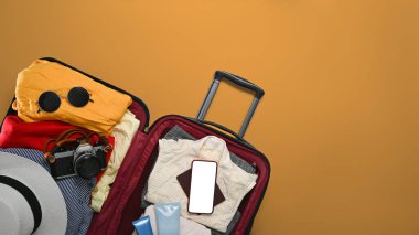 Akıllı telefon, güneş gözlüğü, kamera ve sarı arka planda giysileri olan bavulu aç. Yaz tatiline hazırlanıyorum.
