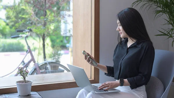 Yan manzara güzel Asyalı kadın koltukta oturuyor ve akıllı telefon kullanıyor.