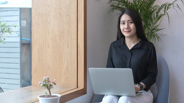 魅力的なアジアの女性起業家は彼女のコーヒーショップでアームチェアに座って、ラップトップコンピュータを使用して — ストック写真