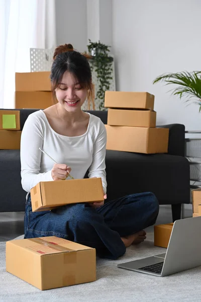 바닥에 앉아 판지 상자 위에 배달용 제품 꾸러미를 준비하고 주소를 쓰고 있는 젊은 아시아 여인을 미소짓는 모습 — 스톡 사진