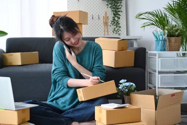 Gülümseyen Asyalı kadın çevrimiçi satıcı cep telefonundan sipariş aldı ve teslimat için paket kutu mal hazırladı
