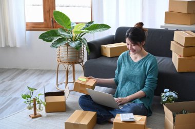 Dizüstü bilgisayar kullanan ve müşterilere teslimat için paket kutu mal hazırlayan gülümseyen genç bir kadın. E-ticaret, Çevrimiçi satış kavramı