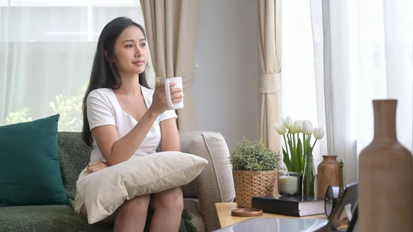 Relaxado millennial asiático mulher sentado no sofá e beber café quente na parte da manhã — Fotografia de Stock