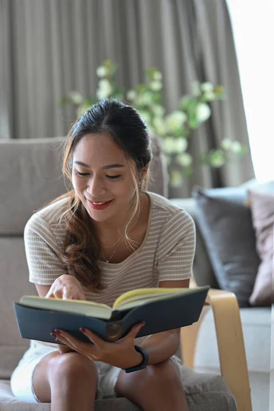 Huzurlu genç Asyalı kadın evinde rahat koltukta dinlenirken kitap okuyor.. — Stok fotoğraf