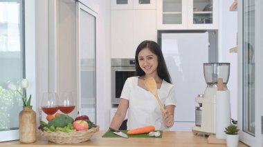 Neşeli Asyalı modern mutfak tezgahında duruyor ve vejetaryen yemekleri yapmak için malzeme hazırlıyor.