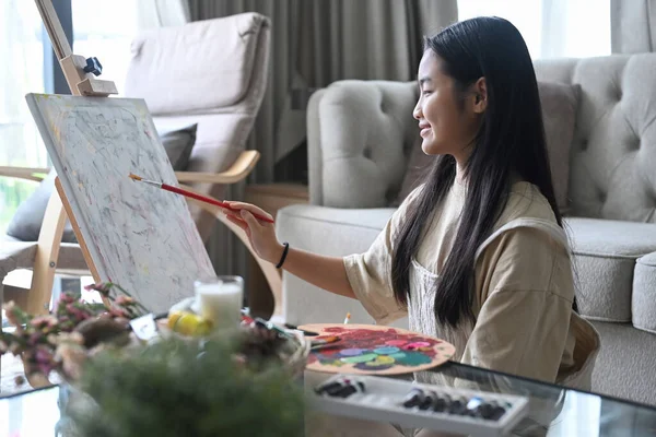 Smiling asian girl painting picture on easel in living room. — Stock Photo, Image