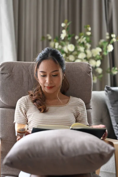 Mujer joven y pacífica leyendo libro en sillón cómodo, disfrutando del tiempo libre en casa. — Foto de Stock