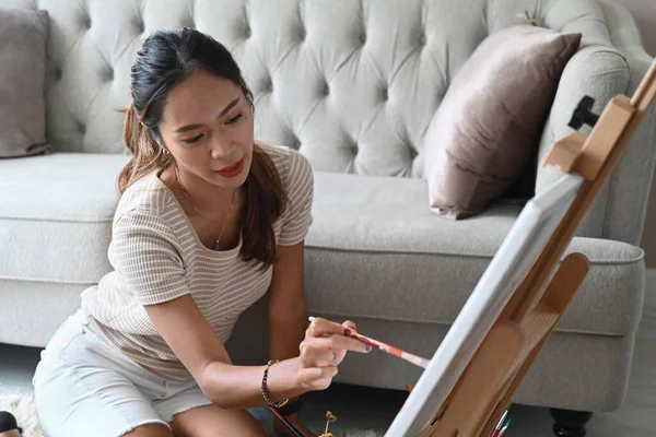 Pretty young woman in casual clothes sitting in living room and painting on easel. — Stock Photo, Image