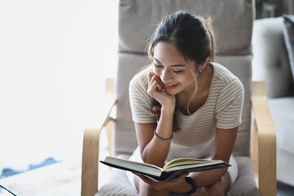 Mujer joven satisfecha leyendo libro en sillón mientras pasa tiempo libre en casa. — Foto de Stock